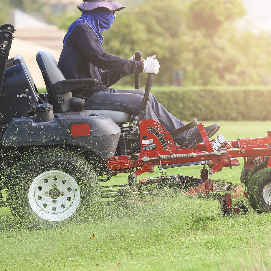 Person mowing grass on a riding lawn mower, ejecting clippings to the side, dressed in protective clothing including a blue long-sleeve shirt and face covering.