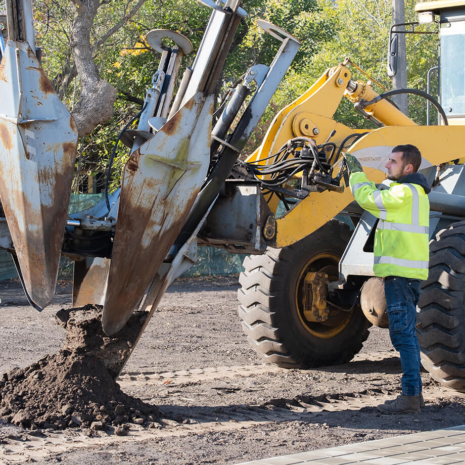 A worker in a high-visibility jacket operates a yellow wheel loader, adjusting the hydraulic arm and bucket in a dirt-covered construction area with trees in the background.