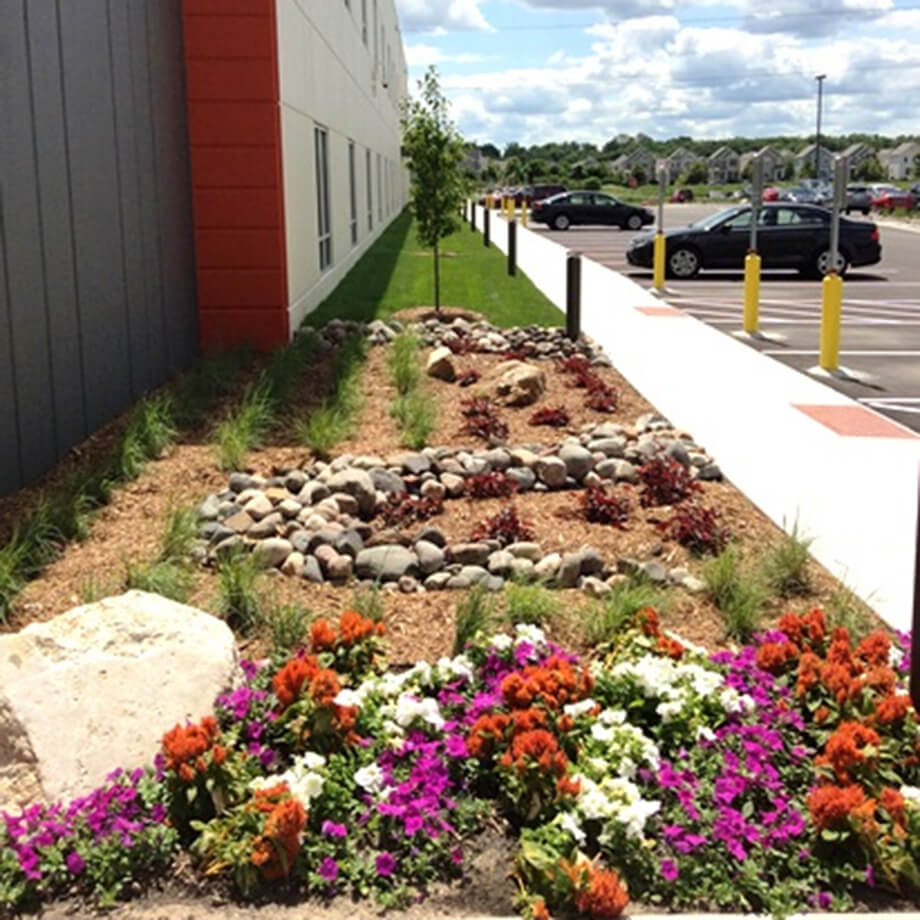A well-manicured landscape bed with vibrant orange and purple flowers, large stones, and mulch alongside a commercial building, under a partly cloudy sky.