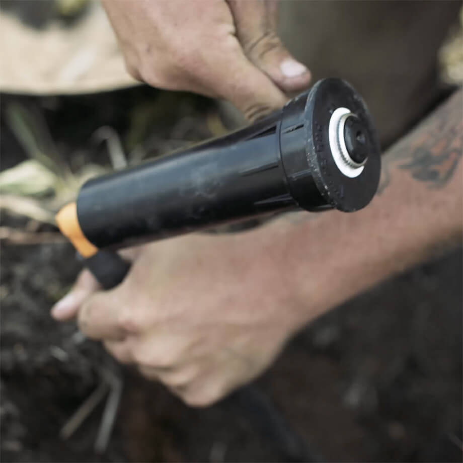 Close-up of a person's hands installing an irrigation system component in the soil, focusing on the black and orange sprinkler head.