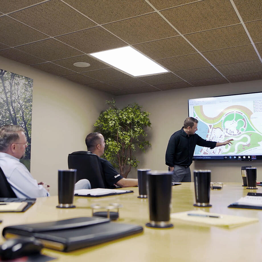 In a conference room, a presenter points to a landscape design displayed on a screen, with three attendees focused on the presentation, a reflection of the company's collaborative planning process.